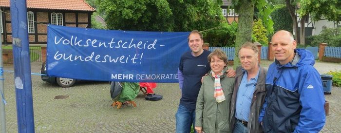 2013-07-03 Ottersberg-Aktion Zwischen zwei Bäumen hängt ein Transparent, mit der Aufschrift "Volksentscheid-bundesweit!". Davor stehen vier Personen und gucken in die Kamera.