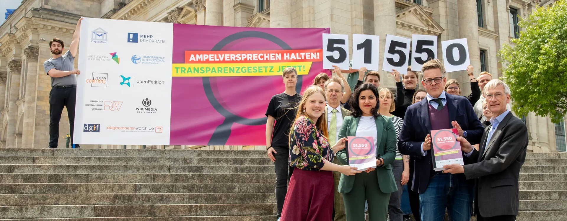 Vor dem Bundestag stehen 16 Menschen, zwei von ihnen halten ein großes Transparent mit der Aufschrift "Ampelversprechen halten! Transparenzgesetz jetzt!", andere halten die Zahl 51550 hoch.