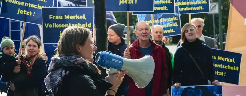 Im Fokus ist ein junge Frau mit einem Megafon und im Hintergrund sind viele Menschen mit Plakaten, welche hochgehalten werden.