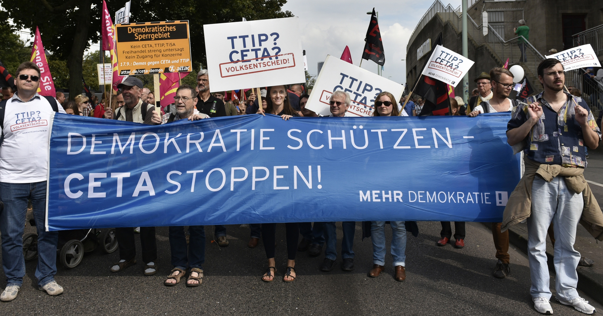 Auf den Bild ist ein Protest mit einem blauen Transparent zu sehen, mit der Aufschrift "DEMOKRATIE SCHÜTZEN-CETA STOPPEN!".