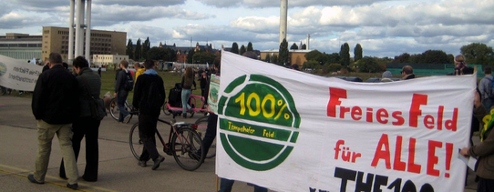 Auf dem Protestmarsch wird ein Transparent gehalten, mit der Aufschrift "100% Tempelhofer Feld" und "Freies Feld für ALLE! www.THF100.de".