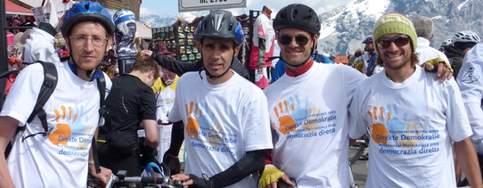 (Kampagne der Initiative zur Sammlung von Unterschriften, September 2013) Vier Menschen sind im Fokus, welche eine weißes T-Shirt tragen, welches direkte Demokratie fordert, während jeder einen Fahrradhelm trägt und sein Fahrrad hält.