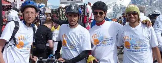 (Kampagne der Initiative zur Sammlung von Unterschriften, September 2013) Vier Menschen sind im Fokus, welche eine weißes T-Shirt tragen, welches direkte Demokratie fordert, während jeder einen Fahrradhelm trägt und sein Fahrrad hält.