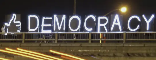 Auf einer Brücke bei Nacht steht in Neon-Schildern "DEMOCRACY" und davor ein Daumen nach oben.