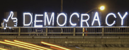 Auf einer Brücke bei Nacht steht in Neon-Schildern "DEMOCRACY" und davor ein Daumen nach oben.