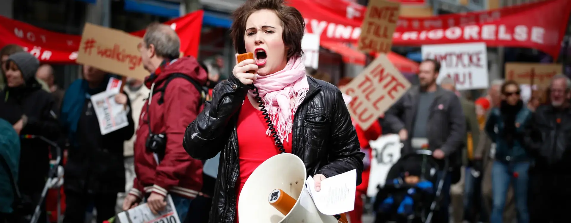 Auf dem Bild ist ein Protest zu sehen. Im Vordergrund steht eine Frau mit einem Megafon und sagt lautstark etwas.