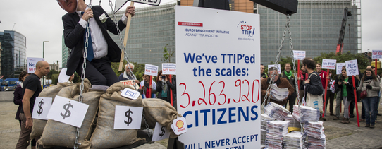 ("Stop TTIP"-Aktion gegen TTIP und CETA vor dem Gebäude der EU-Kommission in Brüssel. | Foto by Alexander Garrido Delgado | Lizenz: CC BY-SA 2.0) Es ist eine altertümliche Waage zu sehen, auf der rechten Seite ist Papier und auf der linken Seite mein Mensch mit ganz vielen Geldsäcken. In der Mitte steht ein Schild, mit dem Text "We've TTIP'ed the scales: 3.263.920 Citizens will never accept TTIP & CETA!".