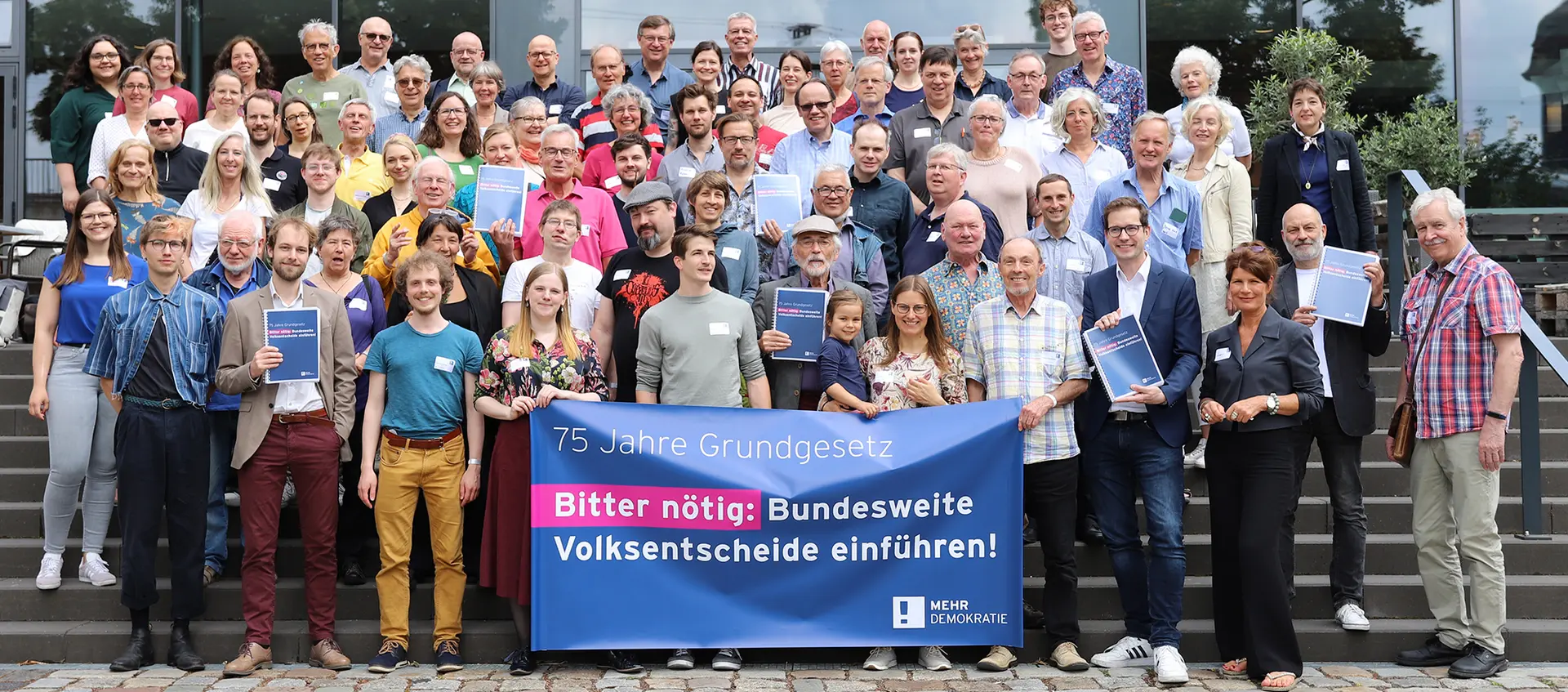 Auf dem Bild stehen ca. 80 Menschen auf eine Treppe verteilt, manche von ihnen halten eine Mappe hoch. Ein Transparent wird hochgehalten mit der Aufschrift: "75 Jahre Grundgesetz Bitter nötig: Bundesweite Volksentscheide einführen", rechts unten in der Ecke das Logo von Mehr Demokratie.