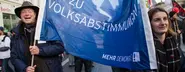 Zwei Menschen tragen ein Transparent auf einem Protest, mit dem Text "JA ZU VOLKSABSTIMMUNGEN".