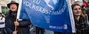 Zwei Menschen tragen ein Transparent auf einem Protest, mit dem Text "JA ZU VOLKSABSTIMMUNGEN".