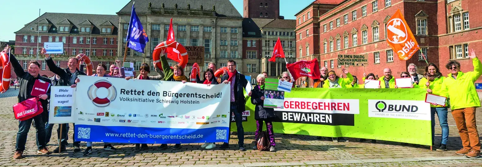 Es stehen circa 30 Menschen mit zwei großen Transparenten auf einem Marktplatz. Auf dem linken Transparent steht "Rettet den Bürgerentscheid!, Absatz Volksinitiative Schleswig Holstein" und auf dem rechten Transparent steht "Volksbegehren bewahren".