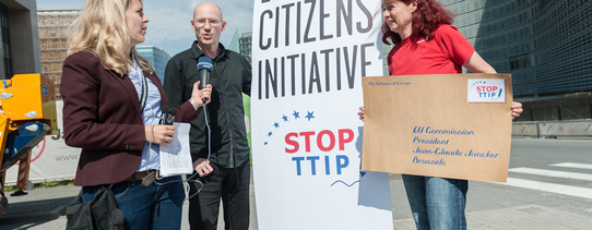 Eine Frau mit einem übergroßen Brief, wo auf der Briefmarke "STOP TTIP" steht, redet mit einer Frau, welche ein Mikrofon in der Hand hat, vor einem zwei Meter großen Transparent, wo "EUROPEAN CITIZENS INITIATIVE STOP TTIP" drauf steht.