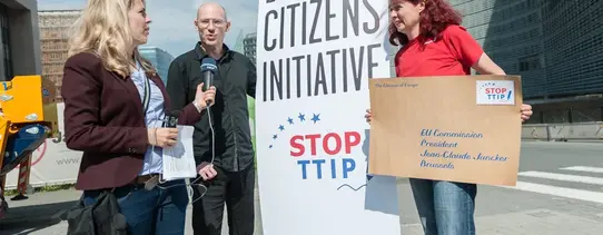 Eine Frau mit einem übergroßen Brief, wo auf der Briefmarke "STOP TTIP" steht, redet mit einer Frau, welche ein Mikrofon in der Hand hat, vor einem zwei Meter großen Transparent, wo "EUROPEAN CITIZENS INITIATIVE STOP TTIP" drauf steht.
