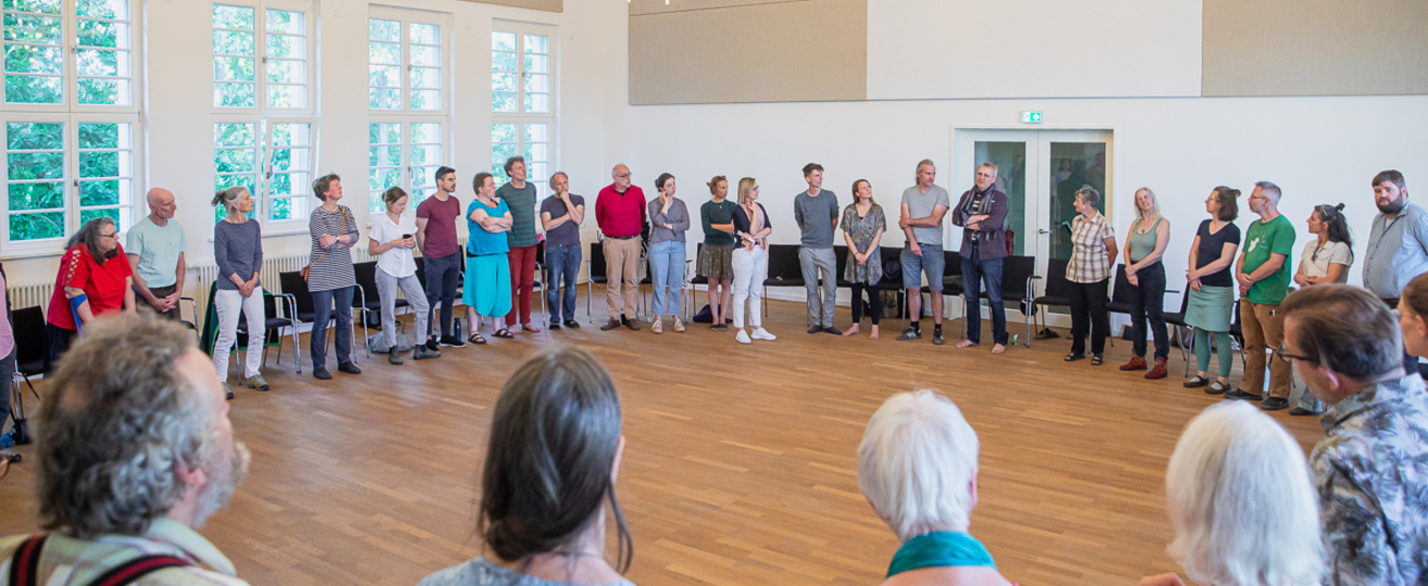 Ein Saal mit 40 Menschen die im Kreis stehen.