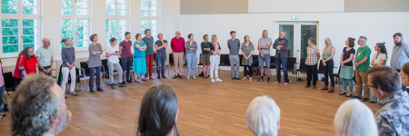 Ungefähr 50 Menschen sitzen in einem Kreis in einem Saal. 