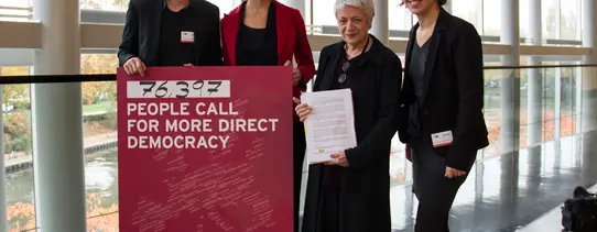 Vier Menschen halten ein Transparent hoch, welches die Aufschrift hat "PEOPLE CALL FOR MORE DIRECT DEMOCRACY".