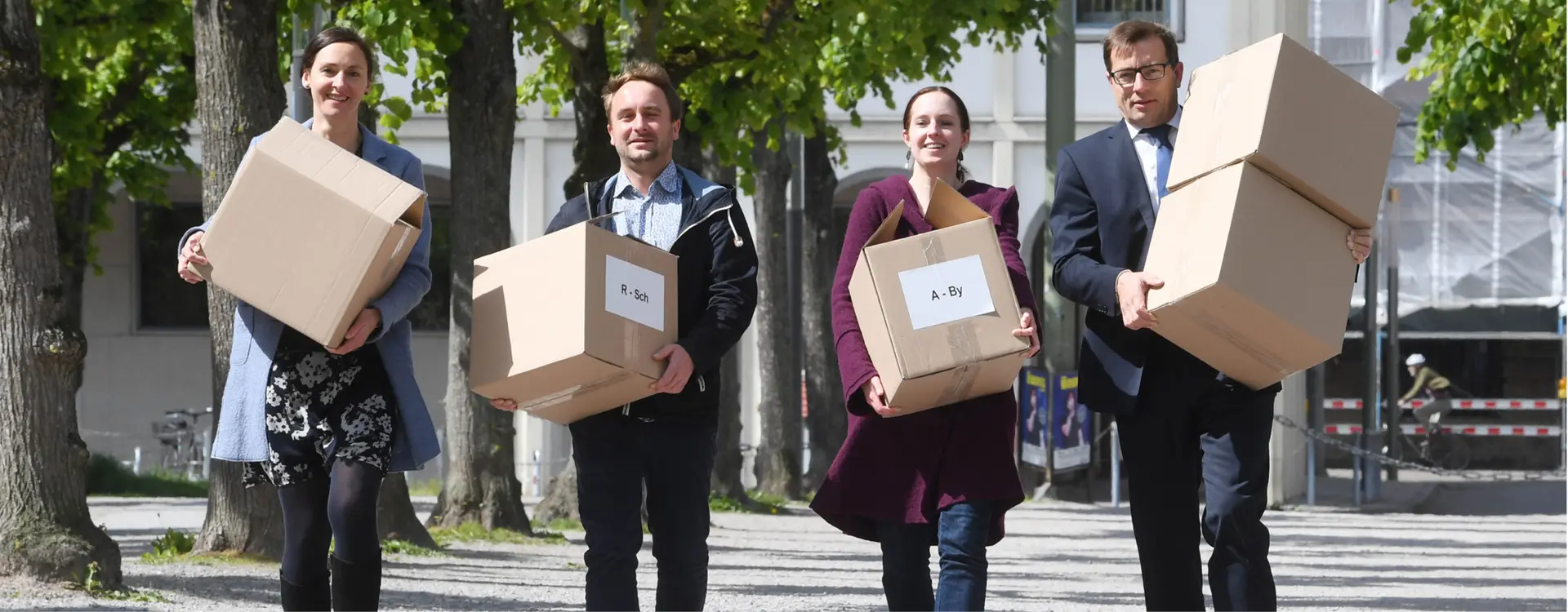 Auf dem Bild sind vier Menschen mit großen Kartons, die in Richtung der Kamera laufen.