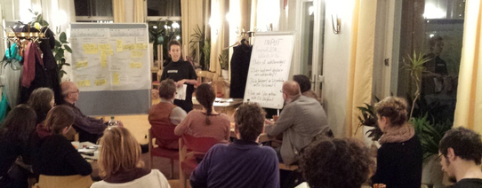 Auf dem Bild sitzen circa 15 Menschen in einer Art Café und vorne ist eine junge Frau, die redet.