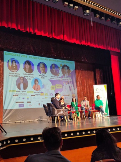 Auf dem Bild ist eine Bühne mit einem Halbkreis von Menschen auf Stühlen zu sehen und im Hintergrund eine Präsentation.