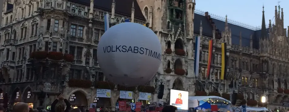 Vor einer Kirche in München stehen hunderte Menschen und im Fokus ist ein weißer Luftballon, mit der Aufschrift "Volksabstimmung".
