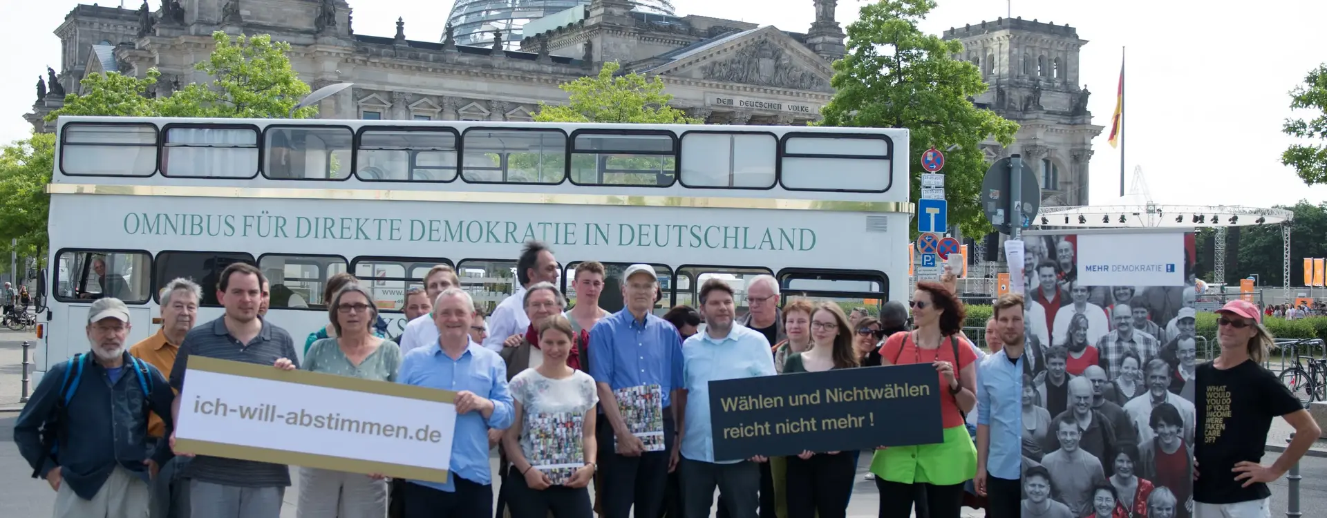 Auf dem Bild stehen Menschen vor dem Bundestag und vor einem Bus, mit der Aufschrift "OMNIBUS FÜR DIREKTE DEMOKRATIE IN DEUTSCHLAND". Die circa 20 Menschen halten Plakate hoch, mit den Texten "ich-will-abstimmen.de" und "Wählen oder Nichtwählen reicht nicht mehr!".