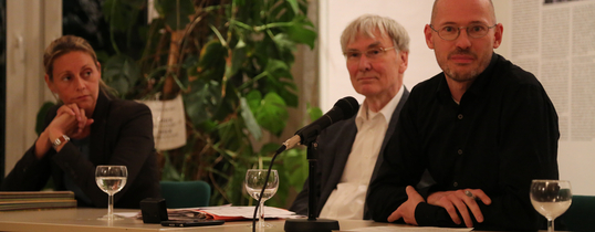 Auf dem Bild sitzen drei Personen vor Mikrofonen an einem Tisch.