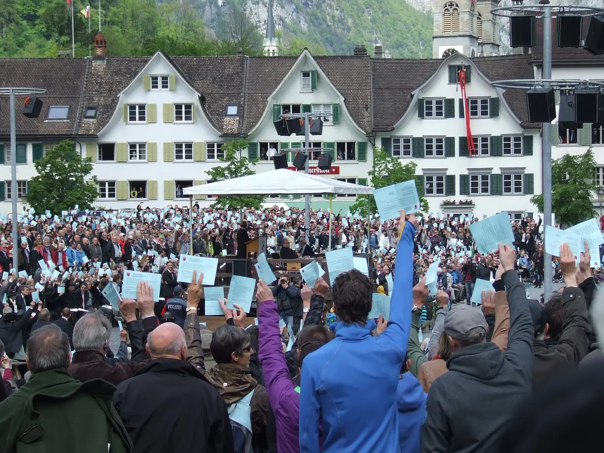 2014-05-09 Schweiz-Reise01 Auf einem Marktplatz stehen mehrere Hundert Menschen und halten einen Zettel in die Luft.
