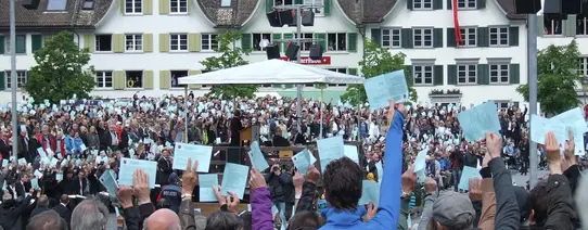 (Schweiz-Reise 2014 | Foto by Cora Pfafferott | Lizenz: CC BY-NC 2.0) Auf einem Marktplatz stehen circa 1.000 Menschen und halten einen Zettel in die Luft.