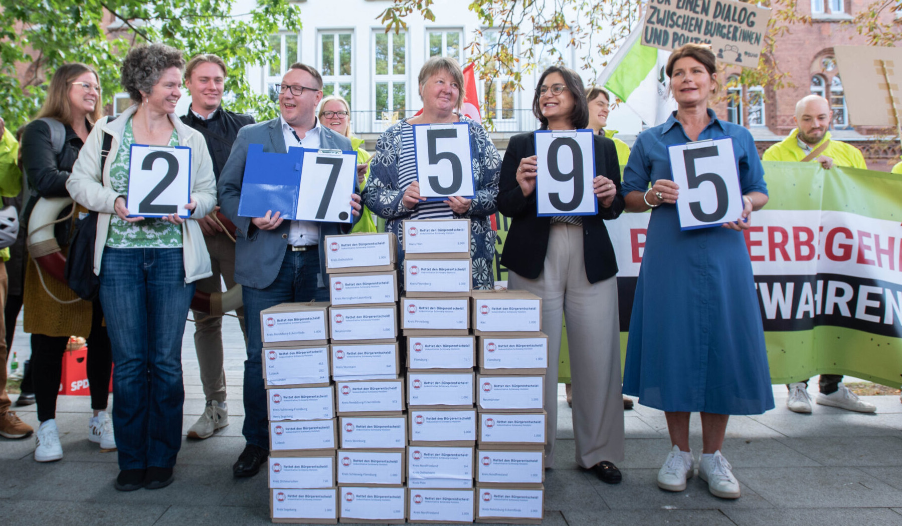 Ein Bild von circa 15 Personen, fünf von ihnen halten die Zahl 27.595 vor einem Haufen Kartons hoch.