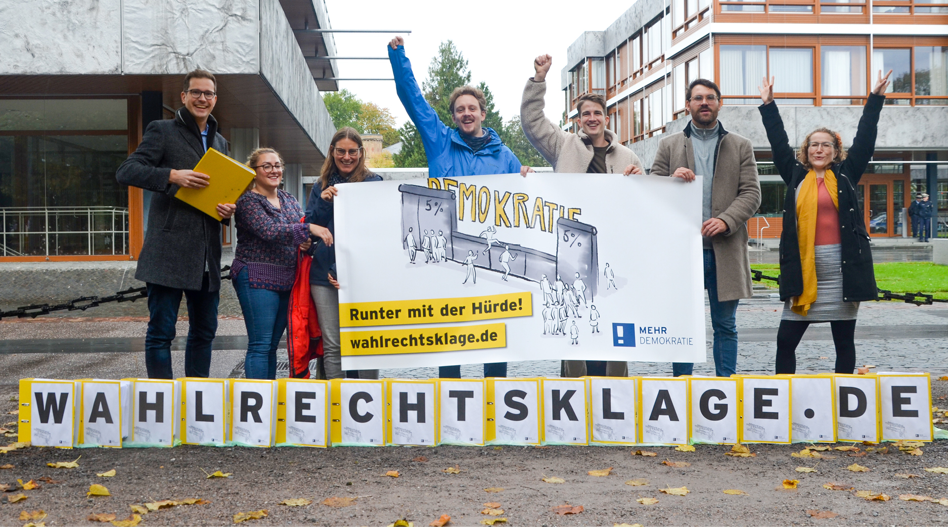 auf dem Bild stehen Aktenordner im Vordergrund. auf jedem ist ein Buchstabe, welche das Wort Wahlrechtsklage.de ergeben. Dahinter stehen sechs Menschen, die sich freuen.