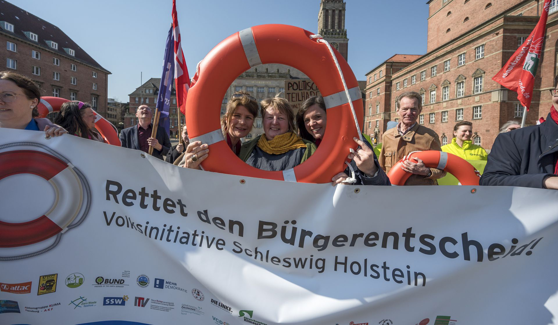 Auf dem Bild ist ein Protest zu sehen. Die Menschen halten ein Transparent hoch mit dem Text "Rettet den Bürgerentscheid!, Absatz Volksinitiative Schleswig Holstein".